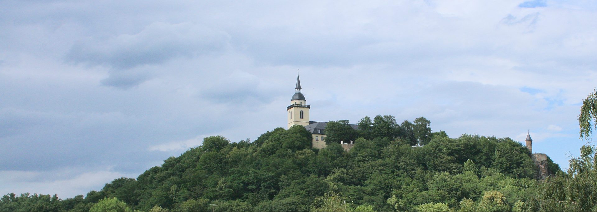 Die barocke Abtei St. Michael mit ihrem Zwiebelturm auf dem bewaldeten Michaelsberg über Siegburg, rechts das Johannistürmchen am Hang unter bewölktem Himmel